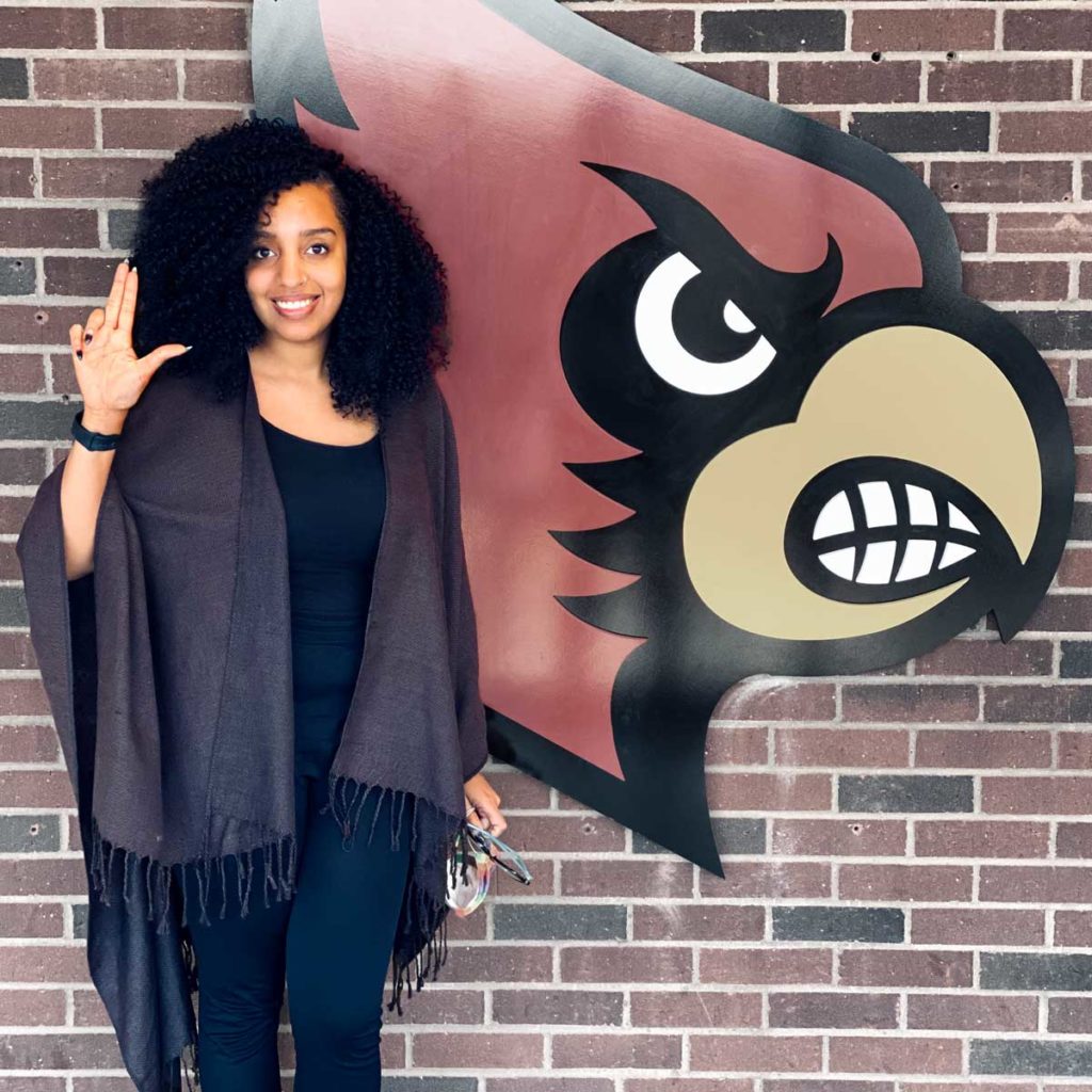 Photo of MIEA Certified Mindfulness teacher Riece Hamilton standing in front of brick wall with University of Louisville graphic. Mindfulness Teacher Certification