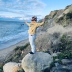 Photo of student Zachary Olivan standing on a rock at the coast with arms outstretched looking up torwards sky and facing ocean with a peaceful look on his face.